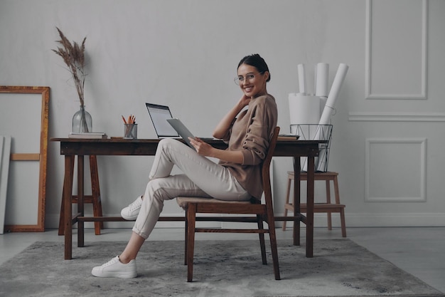 Bella mujer joven sosteniendo una tableta digital y sonriendo mientras está sentada en su lugar de trabajo