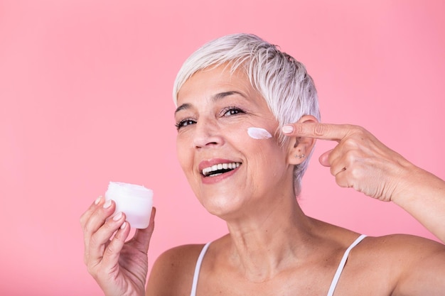 Bella mujer madura sosteniendo un frasco de crema para la piel para la cara y el cuerpo aislada sobre un fondo rosado Mujer anciana feliz aplicando crema hidratante antienvejecimiento y mirando a la cámara Tratamiento anti envejecimiento de belleza