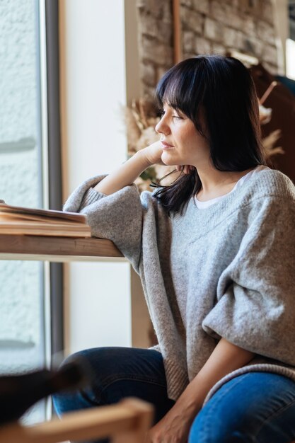 Bella mujer con la mano en la barbilla mirando a otro lado en la cafetería. Imagen de una mujer bonita joven sentada en la mesa de café y libro de lectura.