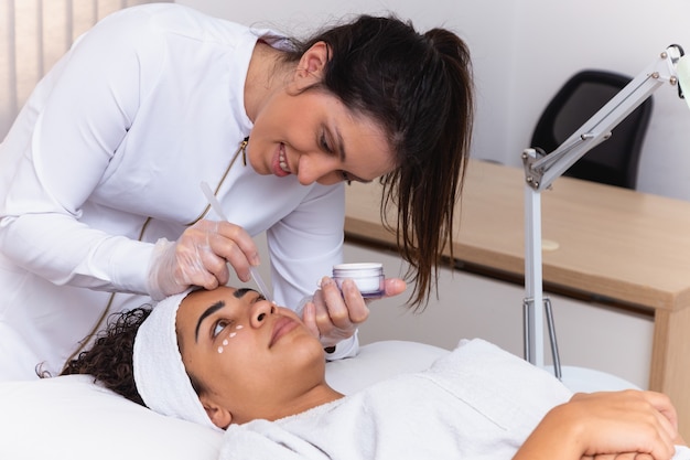 Bella mujer con mascarilla en salón de belleza. Esteticista aplicando crema en la cara del paciente.