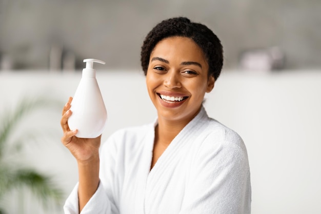 Bella mujer negra sonriente en bata de baño sosteniendo una botella con loción para el cuerpo hidratante