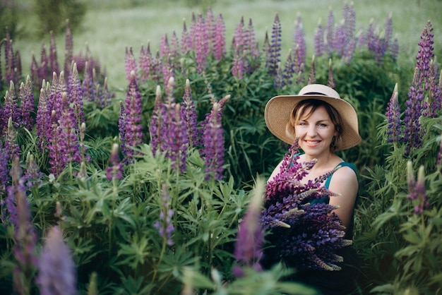 Una bella mujer romántica sonríe con un vestido y un sombrero sentado en un campo de flores con un enorme ramo de flores de lupino púrpura.