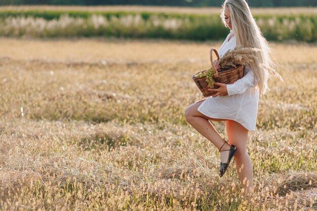 Una bella mujer rubia con el pelo largo en un campo al atardecer lleva una cesta de frutas y un ramo de paja. verano, agricultura, naturaleza y aire fresco en el campo.