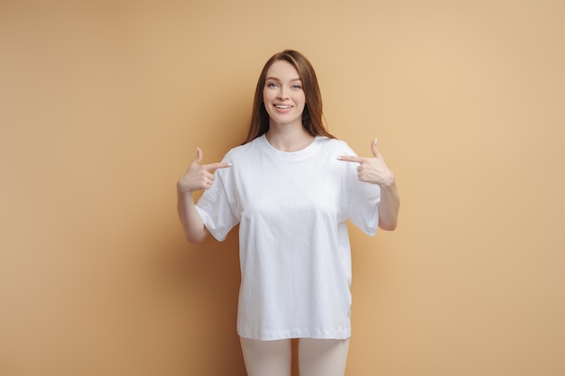 Bella mujer sonriente con camiseta blanca apuntándose a sí misma mirando a la cámara Mockup aislado