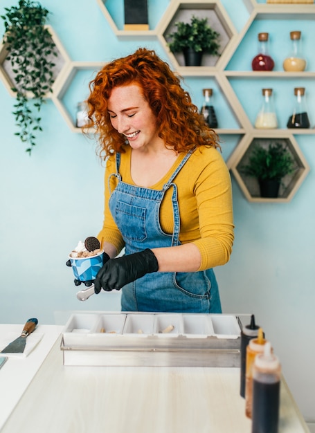 Bella y positiva pelirroja mujer de jengibre sonriendo y trabajando en heladería artesanal. Ella prepara y sirve deliciosos alimentos dulces.