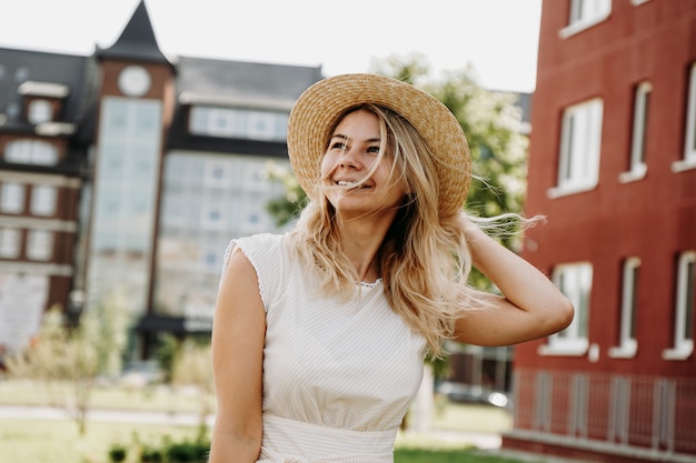 Una bella rubia pasea por una ciudad europea. Mujer con vestido blanco y sombrero de paja, ella sonríe y feliz