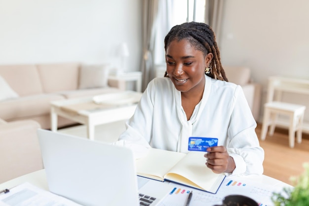 Bella y sonriente joven mujer de negocios africana con una tarjeta de crédito y usando una computadora portátil en la oficina