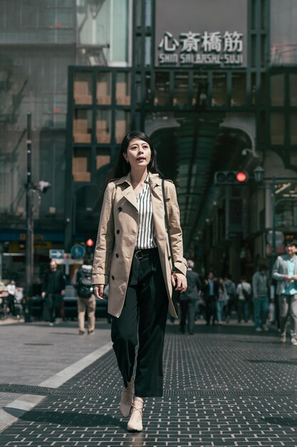 bella trabajadora asiática con abrigo beige con bolsa caminando por la calle. dama de oficina elegante de cuerpo entero que va a trabajar por la mañana en la ciudad de osaka. Traducción en el texto de la pared "área comercial shin sai bashi".