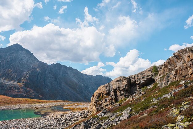 Bella vista de otoño desde las rocas hasta el lago de montaña turquesa contra la alta cima rocosa de la montaña a la luz del sol bajo las nubes en el cielo azul iluminado por el sol hermoso pequeño lago y gran cordillera colores de otoño vívidos