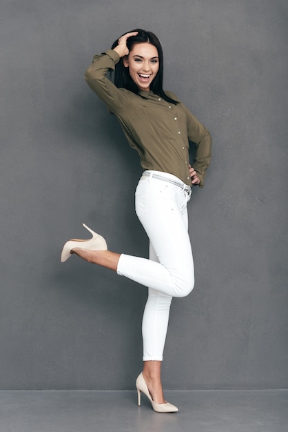 Belleza alegre. Longitud total de mujer joven atractiva en ropa de sport elegante posando sobre fondo gris y mirando feliz