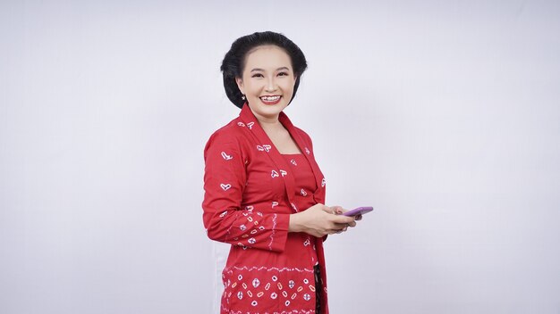 Belleza asiática en kebaya se ríe con smartphone aislado sobre fondo blanco.