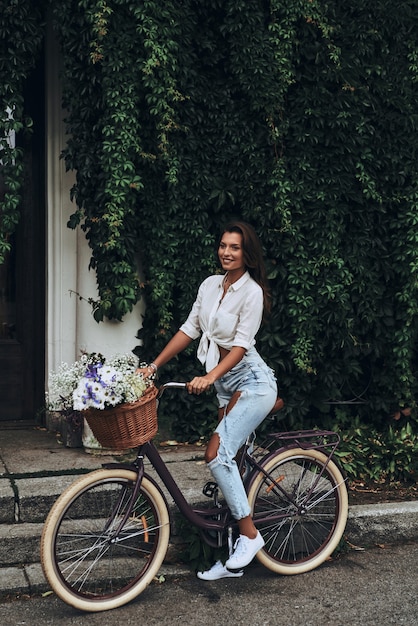 Belleza en bicicleta. Longitud total de mujer joven atractiva en ropa casual sonriendo mientras está de pie frente a la casa cubierta de hiedra