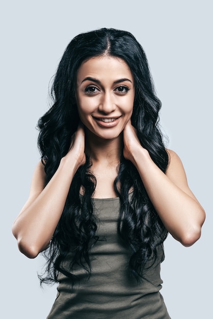 Belleza con cabello curvo. Atractiva mujer joven en ropa casual cogidos de la mano en el cuello y sonriendo mientras está de pie contra el fondo gris