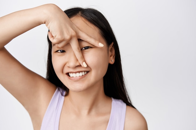 Belleza y cuidado de la piel. Atractiva chica asiática con sonrisa blanca, luciendo feliz y mostrando el signo de la paz en su rostro, de pie en el estudio