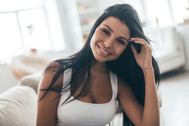 Belleza despreocupada. Retrato de mujer joven atractiva ajustando su cabello y sonriendo mientras está sentado en el sofá en casa