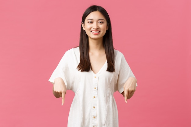 Belleza, emociones de la gente y concepto de ocio y vacaciones de verano. Orgullosa chica asiática feliz y satisfecha en vestido blanco que invita a echar un vistazo a la oferta especial en la tienda, apuntando con el dedo hacia abajo, fondo rosa.