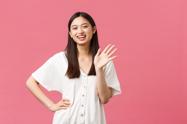 Foto belleza, emociones de la gente y concepto de ocio de verano. amistosa y extrovertida chica asiática bonita con vestido blanco saludando, saludando con la mano y alegre sonriendo, saludando a alguien con gesto de saludo.