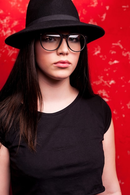 Belleza con estilo. Sesión de estudio de hermosa mujer joven con sombrero y gafas de pie contra el fondo rojo.