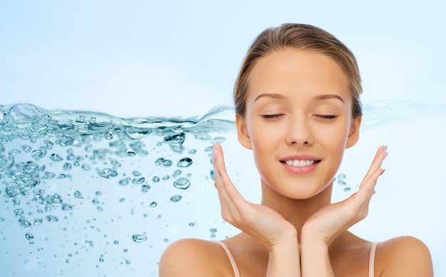 belleza, gente, cuidado de la piel y concepto de salud - cara de mujer joven sonriente y manos sobre salpicaduras de agua sobre fondo azul