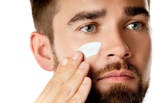 Belleza de los hombres - El hombre joven está aplicando crema hidratante y anti-envejecimiento en el rostro contra el fondo blanco.