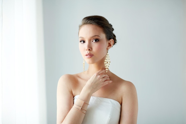 belleza, joyería, accesorios de boda, gente y concepto de lujo - hermosa mujer asiática vestida de blanco o novia con aretes dorados y pulsera