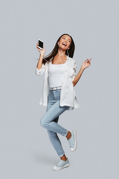 Belleza juguetona. Longitud total de la joven mujer asiática bailando y sonriendo mientras está de pie contra el fondo gris