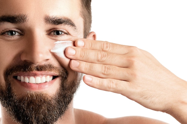 Belleza masculina El joven está aplicando crema hidratante y antienvejecimiento en la cara contra el fondo blanco.