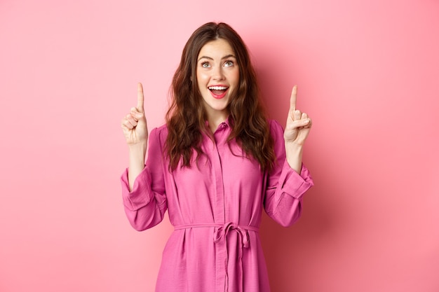 Belleza y moda. Mujer feliz mirando con asombro el espacio de la copia en la parte superior, señalando con el dedo hacia arriba, sonriendo asombrado, mirando la oferta promocional, de pie sobre la pared rosa.