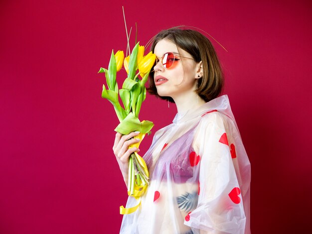 Belleza modelo mujer en impermeable con manojo de flores de primavera. Muchacha hermosa con un ramo de flores amarillas del tulipán. Mujer modelo sorprendida feliz que huele las flores. Regalo del día de la madre. Día de San Valentín.