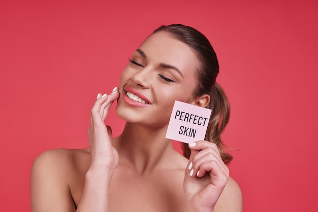 Belleza natural. Hermosa mujer joven sonriendo y tocando su mejilla mientras está de pie contra el fondo rosa