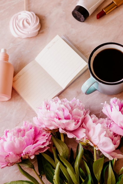 La belleza se posa con un diario, una taza de café, accesorios y peonías sobre un fondo de mármol.