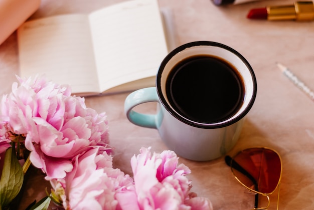 La belleza se posa con un diario, una taza de café, accesorios y peonías sobre un fondo de mármol.