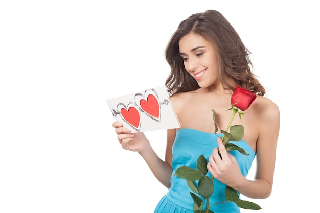 Belleza con tarjeta de San Valentín. Hermosa joven sosteniendo una rosa roja y leyendo una tarjeta de San Valentín mientras está de pie aislado sobre fondo blanco.