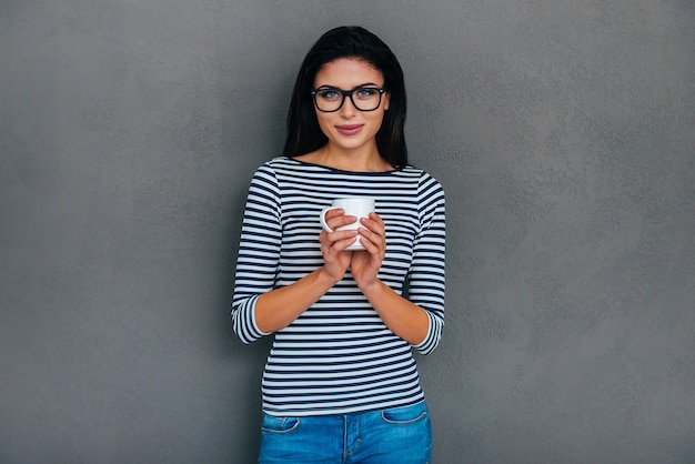 Belleza con taza de café. Atractiva joven mujer sonriente sosteniendo la taza y mirando a la cámara mientras está de pie contra el fondo gris