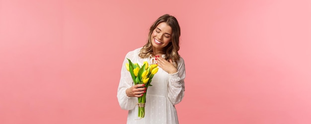 Belleza de vacaciones y concepto de primavera Retrato de una linda y romántica chica rubia complacida recibe un hermoso ramo de flores mirando tulipanes amarillos y toca el corazón sonriendo agradecida