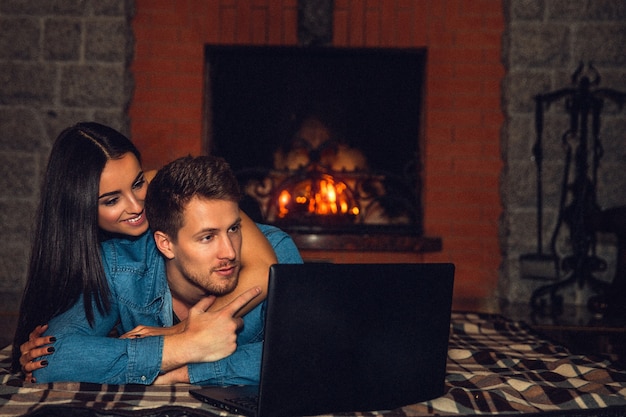 Belo casal jovem deitado no cobertor e assistir filme no laptop. Ela o abraçou por cima e por trás. Eles estão perto da lareira