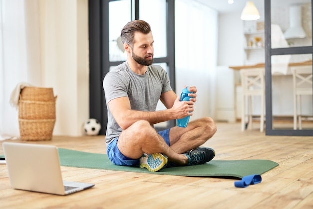 Belo cavalheiro barbudo segurando uma bebida esportiva refrescante enquanto está sentado no chão perto de um laptop moderno