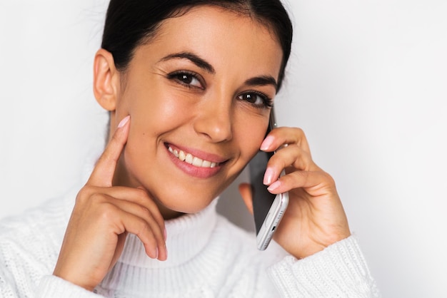 Belo close-up retrato de mulher jovem e atraente feliz com telefone celular com sorriso muito cheio de dentes no fundo branco Linda mulher usando telefone inteligente com cabeça cortada e dedo na bochecha