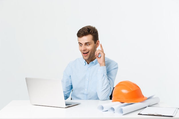 Belo engenheiro está usando um notebook para o trabalho. ele está sentado à mesa e sorrindo. copie o espaço na lateral.