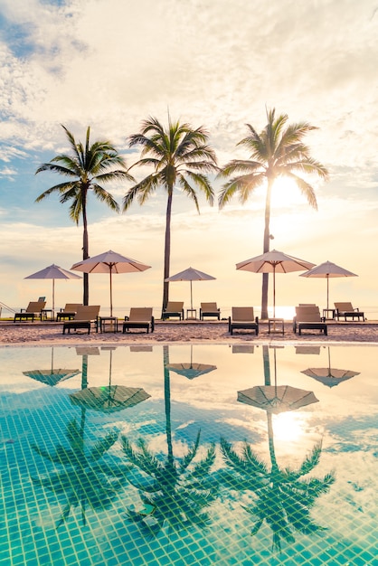 Belo guarda-sol de luxo e cadeira ao redor da piscina ao ar livre do hotel e resort com coqueiro no céu do pôr do sol ou amanhecer
