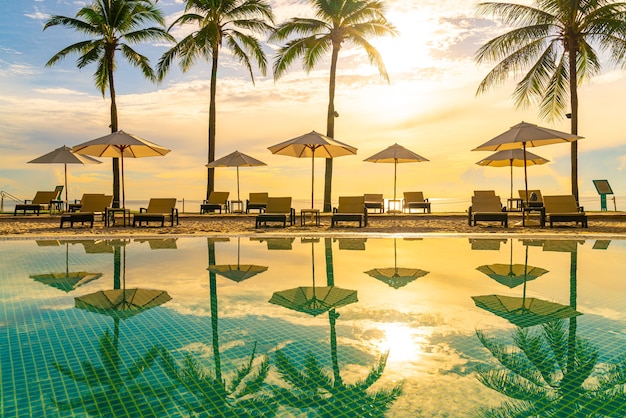 Belo guarda-sol de luxo e cadeira ao redor da piscina externa do hotel e resort com coqueiro no céu do pôr do sol