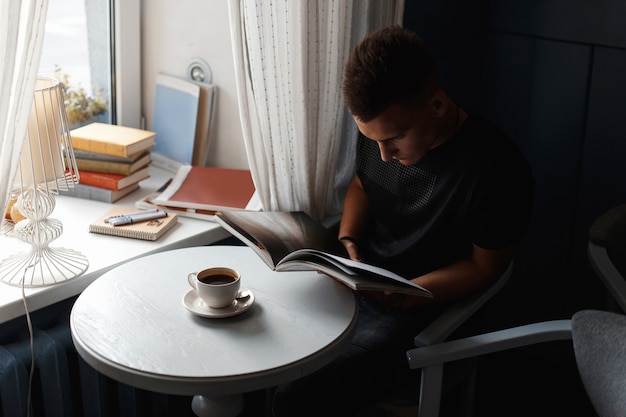 Belo homem moderno descansa em um restaurante, lendo um livro e bebendo café.