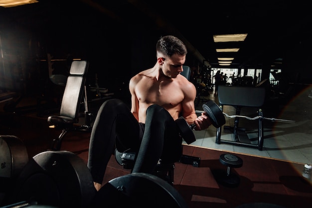 Belo homem musculoso se exercitando com halteres pesados em um ginásio moderno e escuro