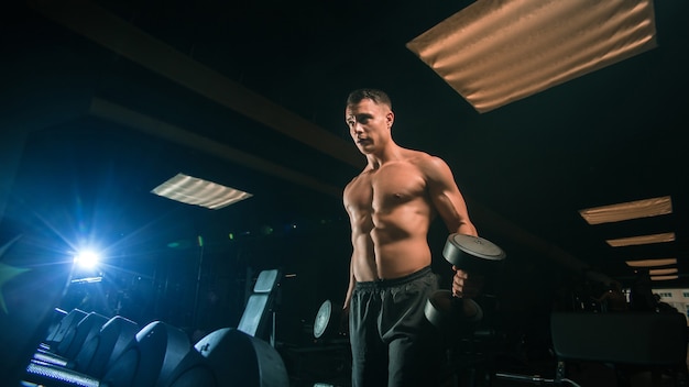 Belo homem musculoso se exercitando com halteres pesados em um ginásio moderno e escuro