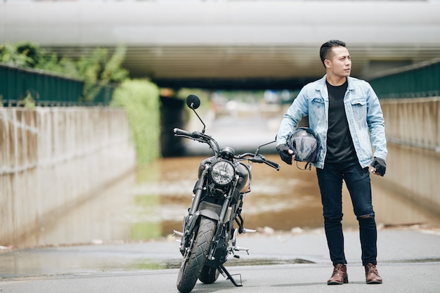 Belo jovem asiático sério em jaqueta jeans, ao lado de sua motocicleta com um capacete nas mãos