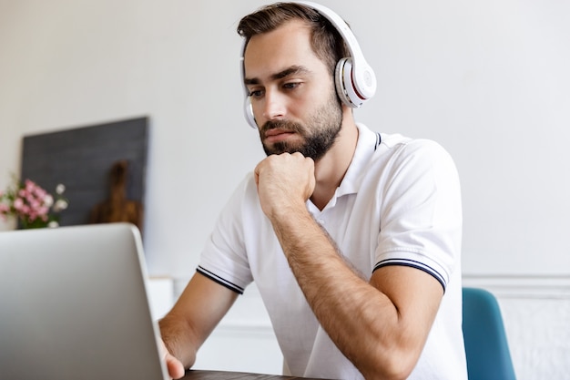 Belo jovem barbudo sério sentado à mesa em casa, usando um laptop