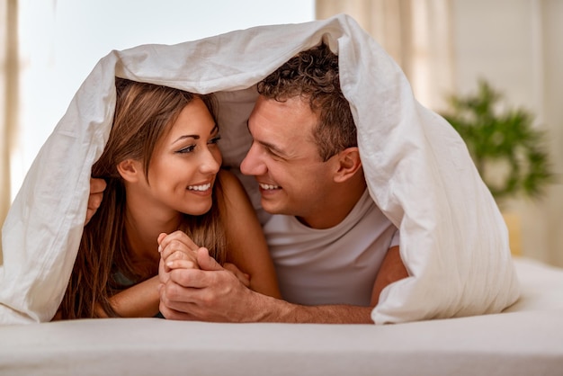 Foto belo jovem casal apaixonado olhando um ao outro com amor e tendo uma manhã preguiçosa na cama coberta com cobertor.