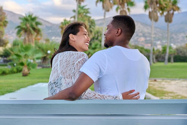 Belo jovem casal multicultural sentados juntos em um banco em um parque tropical