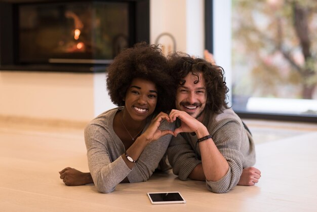 belo jovem casal multiétnico com computador tablet mostrando um coração com as mãos no chão em frente à lareira no dia de outono