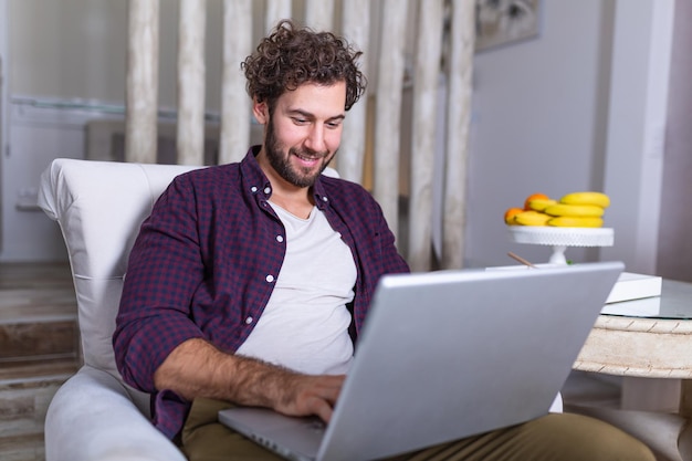 Belo jovem relaxando em casa na poltrona, sentado na sala de estar com o laptop, sorrindo. Digitando uma nova postagem no blog. homem bonito usando laptop com um sorriso enquanto está sentado no sofá em casa